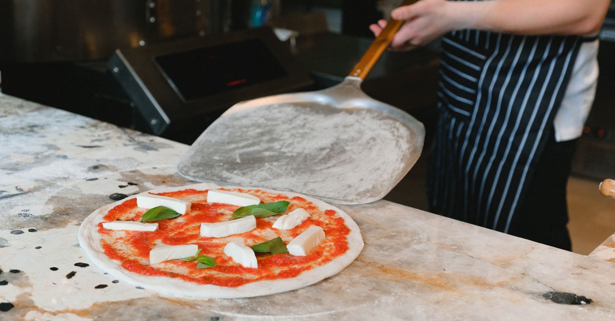 A chef prepares uncooked pizza with cheese and basil on a marble counter.